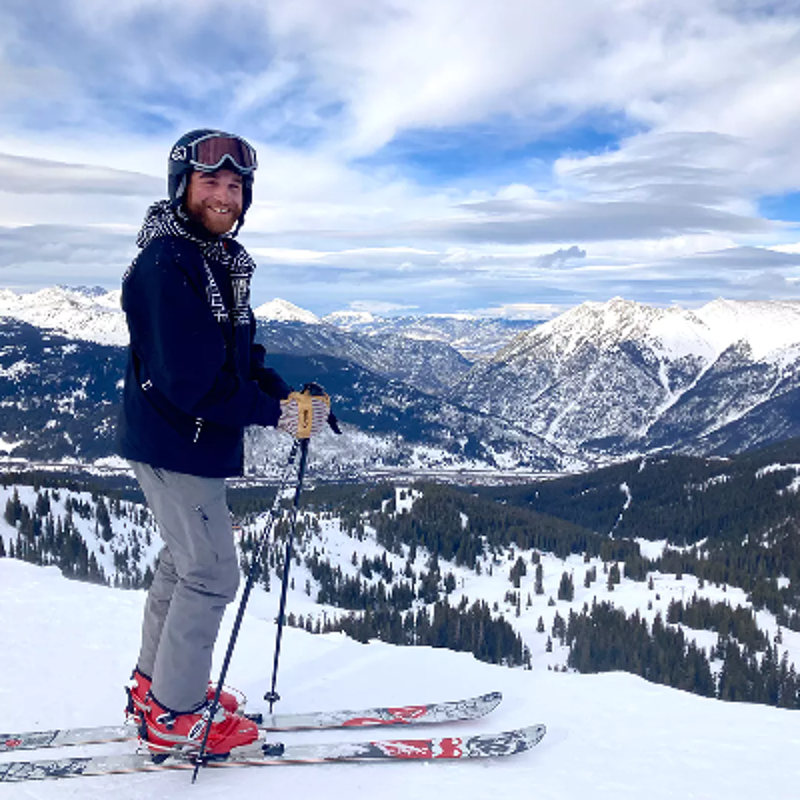 The image shows a man on skis, smiling at the camera. He is wearing a helmet, goggles, and winter clothing. The background features a snowy mountain range under a cloudy sky. The man appears to be enjoying a day of skiing.